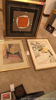 Photo showing three framed French-themed art pieces on the floor, two with orange square and white figures, one with vintage ad style.