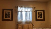 Photo showing both framed watercolor paintings hanging on a wall with curtains and towels below.