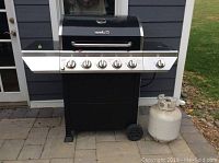 Full frontal photo showing the black and silver Nexgrill 5-burner propane outdoor grill with attached propane tank on the right side, positioned on a stone tile surface in front of a building.