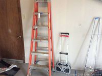 Six-foot orange Louisville step ladder next to silver and black wheeled folding Magna Cart against a wall