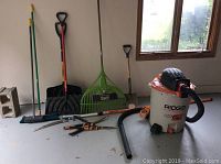 Full view of shop vac and yard tools including shovels, rakes, pruners, hedge trimmers, and broom.
