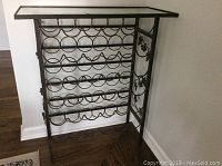 Front view of metal framed wine rack with glass top, showing rows of circular metal slots for wine bottles and decorative side detail with grape and leaf accents.