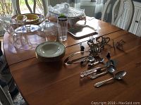 Full table view showing white Marinex serving dish with lid, various Lenox bowls stacked, pie dish, and assortment of silver serving utensils and spoons laid out