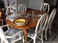 Oval wooden dining table with six white wooden chairs with blue patterned seats, various glass and kitchenware items placed on table.