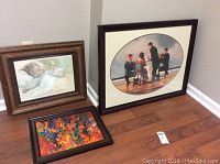 Three framed art pieces on the floor: print of sleeping child, print of men around a table on a boat, and colorful abstract painting