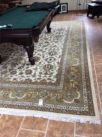 Photo showing most of one side of the rug under a pool table on tiled floor, highlighting the cream center field and olive green border with floral motifs.