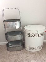 White metal laundry hamper with patterned cut-out bands and hinged lid beside three-tier galvanized metal storage rack