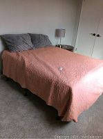 Full view of bed made with coral pink quilted bedspread and two gray pillows on metal bed frame.