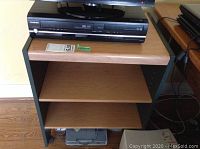 Front view of faux wood storage stand with two shelves and a top holding electronic devices (contents not included).