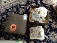 Photo showing closed tureen with floral and leaf painted design and yellow knob, cheese dish with floral motif and ridged sides, and a box on patterned carpet.