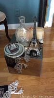 Photo showing the mirrored stand surface with various decorative items including glass decanter, metal Eiffel Tower model, circular metal tray and small wooden boat ornament.