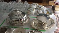 Silver plated serving pieces displayed on a table including a large domed cover, footed tray, and chafing dish alongside porcelain dishes.