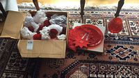 Cardboard box filled with red glassware items, including cups wrapped in protective foam, set on patterned rug.