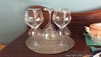 Six clear wine glasses arranged on round silver-tone beaded mat on wooden surface