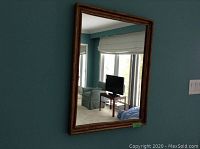 Rectangular wall mirror mounted on blue-green wall reflecting part of a living room with window and TV.