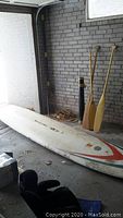 Photo showing the Mistral Surfer paddleboard and three wooden paddles against a brick wall in a garage setting. The board shows signs of wear.
