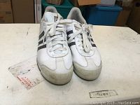 Front view of pair of Adidas Samoa sneakers showing white color, black stripes and scuffed toe areas.