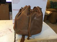 Brown satchel shoulder bag with leather drawstring closure and chain leather combination strap shown on table with background of a chair and boxes.