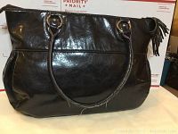 Front side of large black laptop bag showing handles attached with metal rings and smooth shiny faux leather surface.