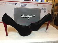 Side view of Herstyle black faux velvet platform heels with red heel against a gray box in the background