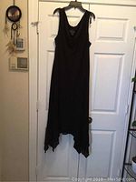 Front view of black dress hanging on white door showing sleeveless design and asymmetrical hem.