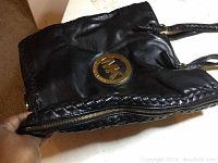 Side view of black leather tote showing braided edge and side zipper partially opened to show expansion capability and gold MK logo.