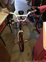 Front view of pink children's bike with white handlebar and pink grips, black padded seat, training wheels and plastic pegs on front wheel.