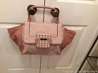 Photo of Guess mauve and pink handbag hanging on doorknobs, showing front, flap, and handles with stud details on clasp.