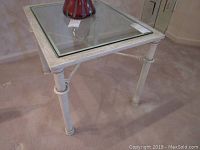 Side table full view showing square glass top set into a painted metal frame with neoclassical detail legs and cross supports.
