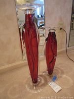 Two tall cranberry and clear glass candle stands, one taller measuring 22 inches, placed on carpeted floor in front of mirror.