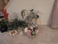 Photo showing various silk flowers arranged in gold-tone vases, two large ornate metal candle holders, green artificial trailing plant, ceramic flower-shaped dish, and a small white rectangular item on carpet.