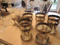 Photo of nine clear highball glasses with silver bands and monogrammed "M", along with tall etched stemmed glasses and some other clear glassware with cut patterns.