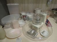 Photo showing various kitchen items: ceramic mugs, plastic containers, a food chopper, a ceramic pitcher, and kitchen utensils arranged on a surface.