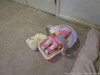 Bitty Baby doll seated in pink and purple carrier with flower teddy bear beside it on carpet.
