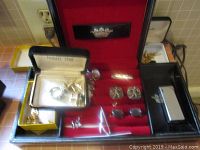Top-down photo of black display box open to reveal cufflinks and other accessories arranged on red velvet, also several boxes lined up with gold and silver-tone cufflinks and a silver-tone money clip. Some cufflinks have jade stones and sword-shaped pins are visible.