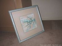 Framed mixed media artwork leaning against a wall on carpet, showing the full piece including silver frame and beige mat.