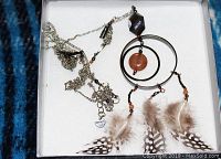 Close-up photo of necklace showing the unique silver-tone chain, circular pendant with agate and hematite stones, and hanging rooster feathers inside the box.