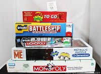 Five boxed games stacked, including Apples To Apples To Go, U-Build Battleship, U-Build Monopoly, Despicable Me metallic puzzle, and vintage Monopoly box.