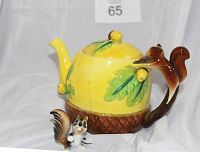 Front-left angled view of a bright yellow acorn-shaped porcelain teapot with green leaves and acorn decorations. Squirrel-shaped handle in brown, and separate porcelain squirrel figurine at base.