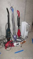 Front view showing three vacuum cleaners including Dirt Devil handheld, Shark upright, Hoover upright, along with DeLonghi heater and two irons laid on the floor.
