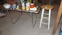 Folding table with light-colored laminate top and metal legs with wear, wooden kitchen stool with round seat and white painted legs, placed on concrete floor.