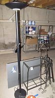 Black metal floor lamp, metal aquarium stand, two mirror panels, set of metal bed posts, and metal wall shelf frame arranged on basement floor with exposed ceiling beams and concrete walls.