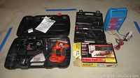 Three cordless power tools with chargers and boxes laid out on floor: Black and Decker multi tool in black case, Craftsman rotary tool in box with attachments, Craftsman cordless screwdriver with charger and blue box of bits.