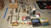 Photo of various hand tools and hardware spread on the floor including clamps, hammer, files, trowel, box with screws, and tool racks