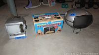 Three vintage items on basement floor: left is silver Coby portable TV with dimensions 8x8x7.5 inches noted; center is boxed Radio Shack TV Scoreboard; right is silver globe-shaped Panasonic portable TV.