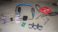 Workout equipment including Blue and black Ab Sculptor, Red Fitness DX bench, green hand weights, black ab roller, and manual on concrete floor.