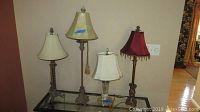 Four table lamps arranged on a glass table showing various lamp shades and metal and glass lamp bases.