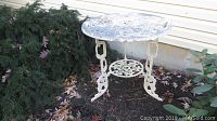 Outdoor metal table with ornate floral top and curved legs showing wear and paint loss, positioned beside shrubbery near house siding.