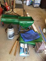 Photo shows two green Scotts Speedy Green lawn spreaders, a white Craftsman 2-gallon hand sprayer with black hose, and two tarps (blue and green) laid out on a garage floor with other garden-related items visible.