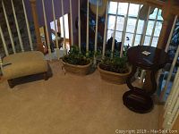Photo showing footstool, two live plants in plastic planters, and dark wood side table next to a railing.